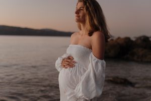 A pregnant woman in a white off-shoulder dress stands by a serene lake at sunset.