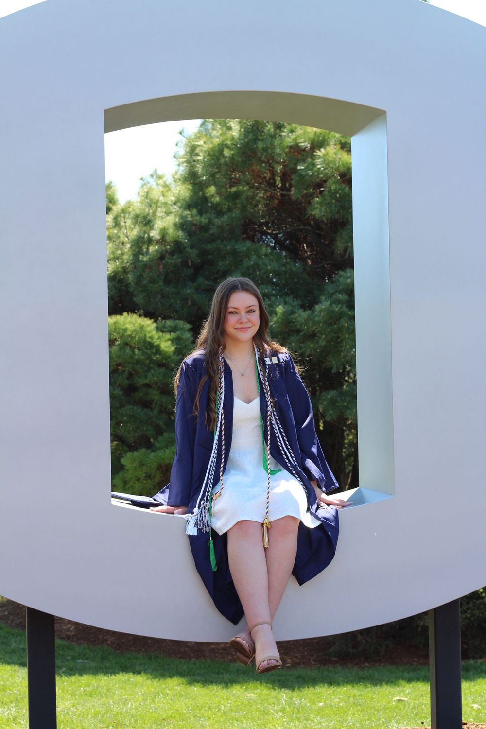 A person in a navy blue jacket and white dress poses creatively in a curved architectural structure outdoors.