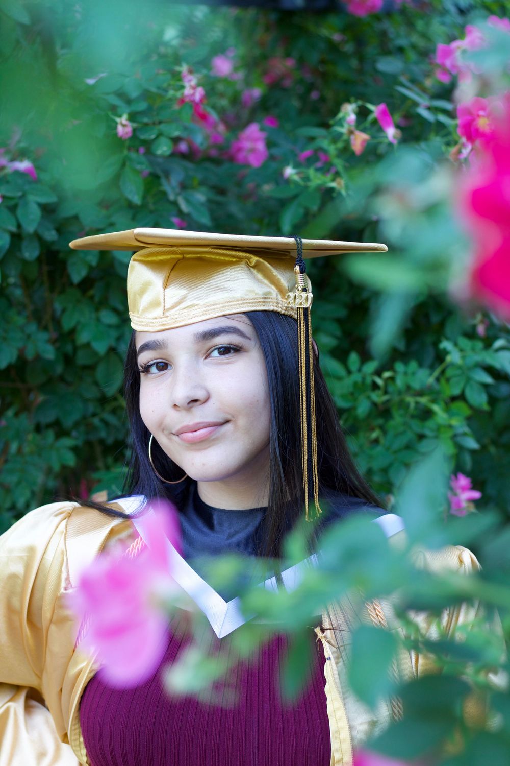 A graduation portrait with gold cap and gown framed by pink flowers and green foliage in a garden setting.