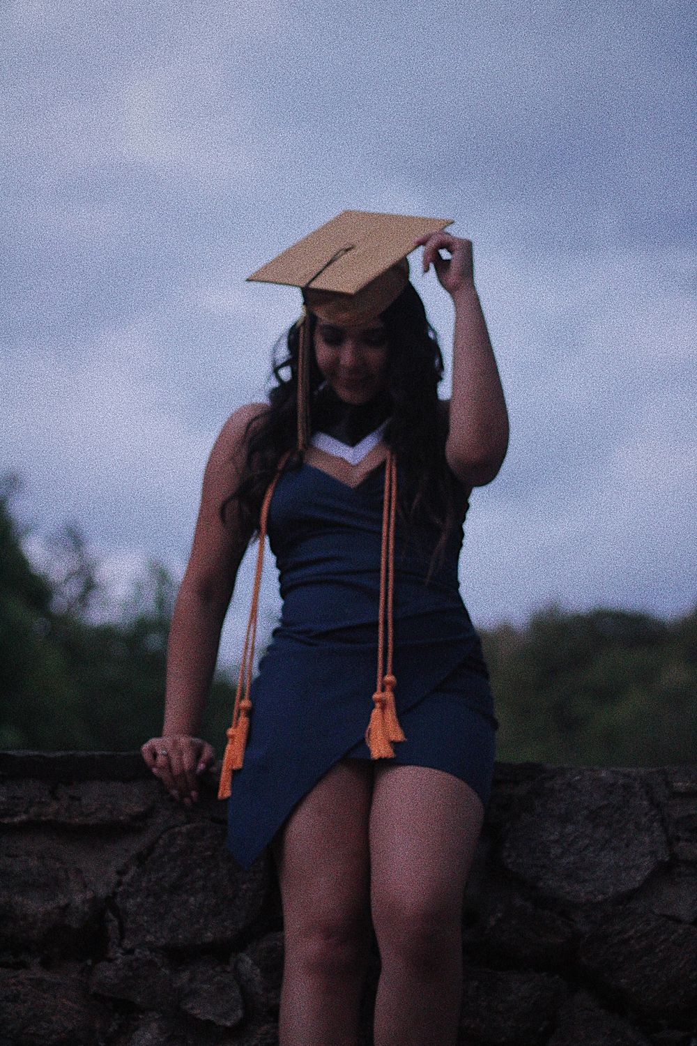 A graduation photoshoot series showing someone in a navy blue dress and graduation cap posing against a cloudy sky.
