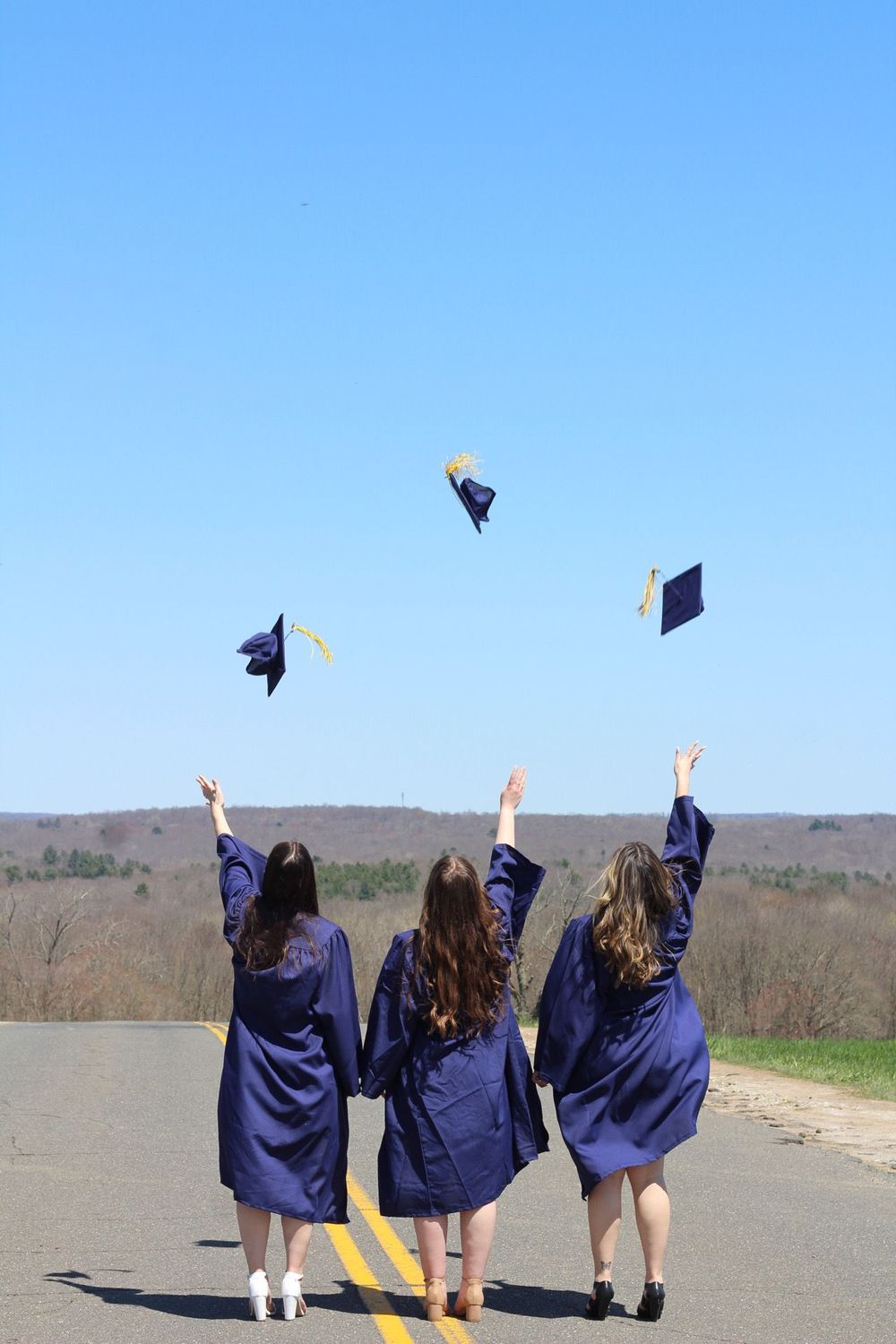 Three graduates in blue caps and gowns throw their caps in the air while celebrating on a rural road.