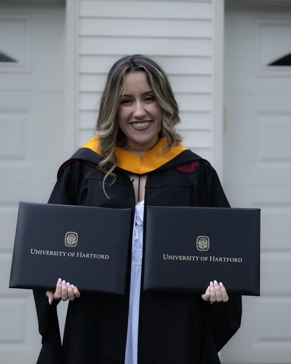 Graduates holding University of Hartford diploma covers while wearing black caps and gowns with gold stoles.