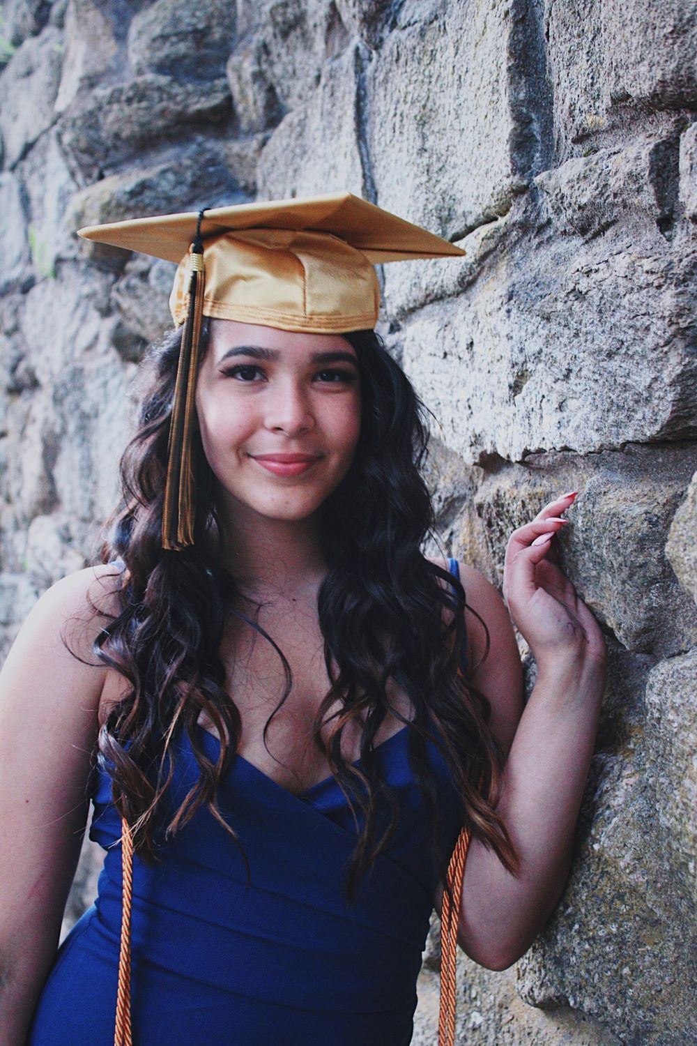 Graduate in blue dress and gold cap poses against stone wall in series of casual graduation photos.