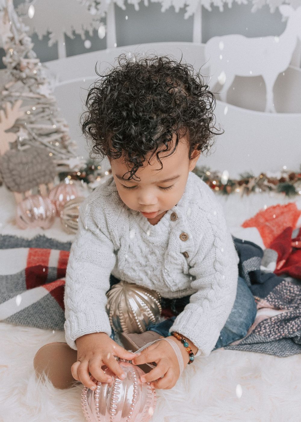 A child in a white cable knit sweater plays with holiday decorations and ornaments in a festive winter setting.