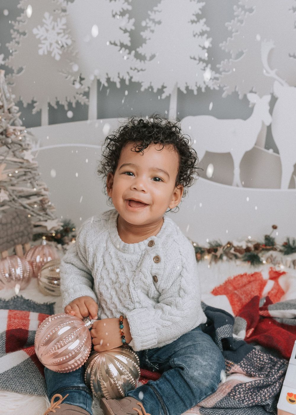 A child in a white sweater and jeans plays with silver and rose gold Christmas ornaments during a winter photoshoot.