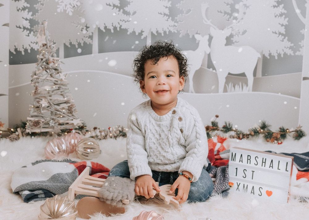 A toddler sits on a white floor surrounded by Christmas decorations and silver tinsel trees in a festive holiday setting.