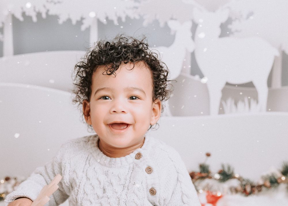 A baby wearing a white sweater smiles and plays in a bright, festive holiday setting with white decorations.