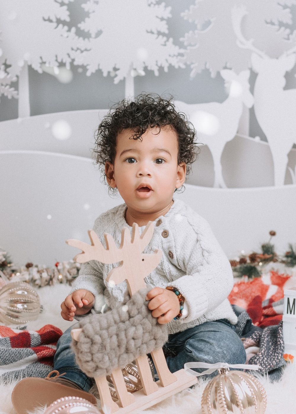 Baby in white sweater and jeans plays with gray and white winter-themed decorations in a festive setting.