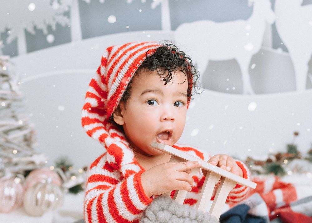 A baby in a red and white striped holiday outfit plays with Christmas decorations in a winter scene.