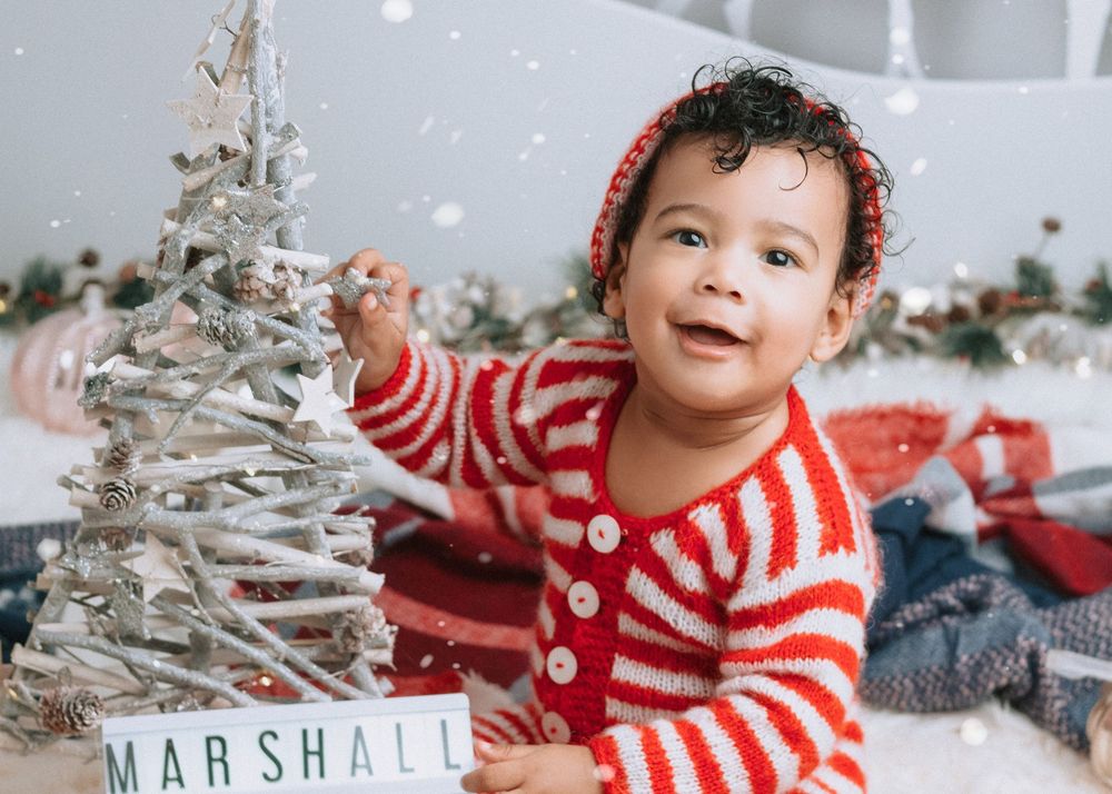 A child wearing a red and white striped sweater sits among Christmas decorations and holiday decor.