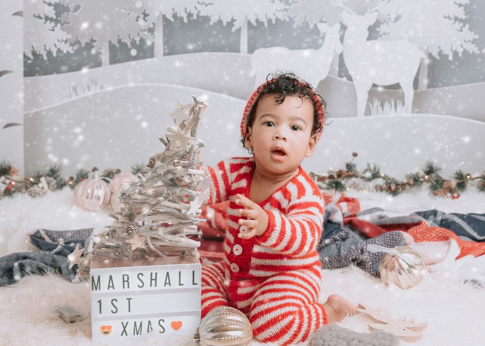 A baby in red and white stripes sits on a snowy winter backdrop with Christmas decorations and festive props.