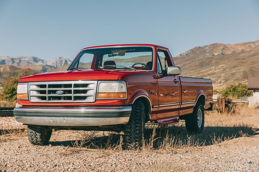 A series of vintage Ford pickup trucks parked in a desert landscape with mountains in the background.