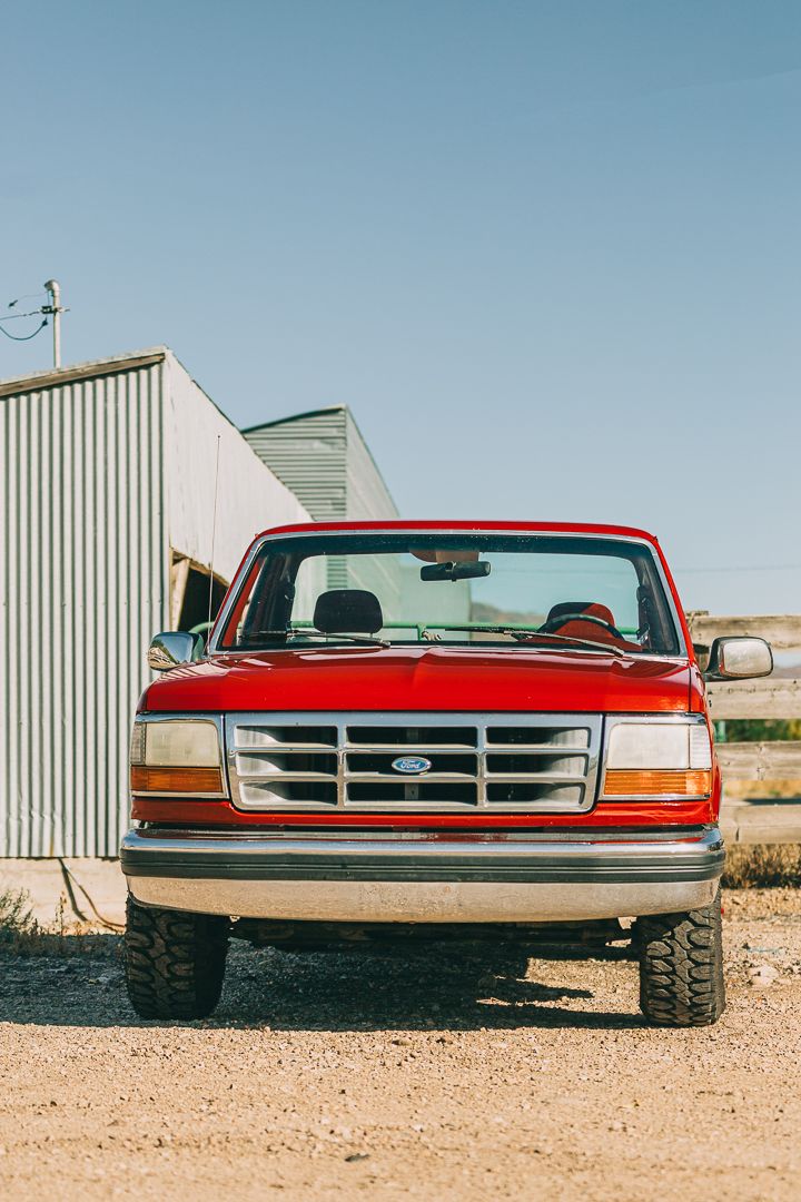Head-on view of a bright red vintage Ford F-150 pickup truck parked in front of metal buildings.
