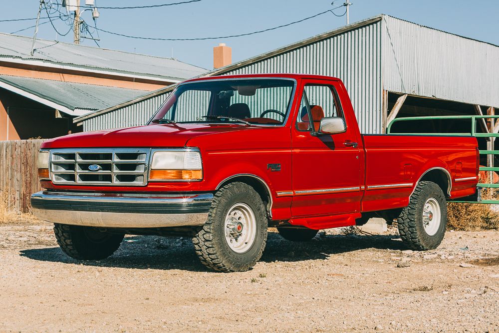 Classic red Ford F-150 pickup truck from the 1990s parked on dirt ground near industrial buildings.