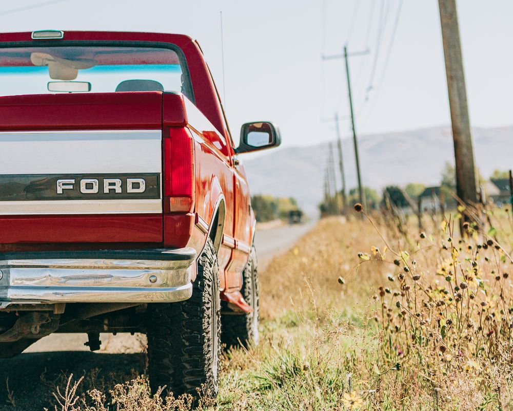 Red Ford pickup truck parked on rural road with utility poles and dry grass in background.