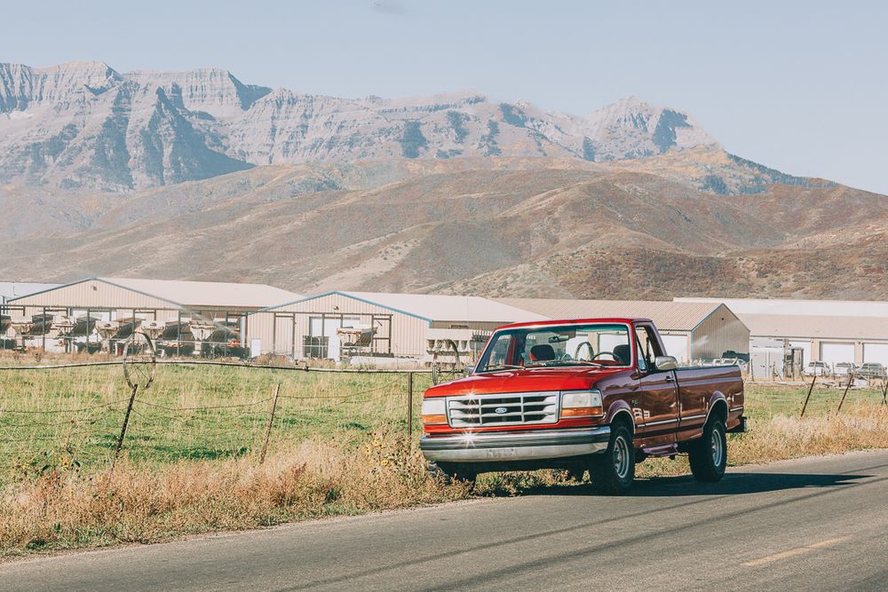 Panoramic view of desert mountains with red pickup truck in foreground against rugged landscape.