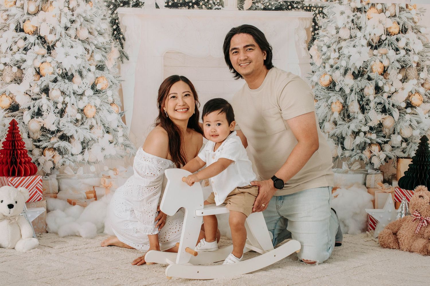 Family poses together in matching white outfits for holiday photos in front of decorated Christmas trees.