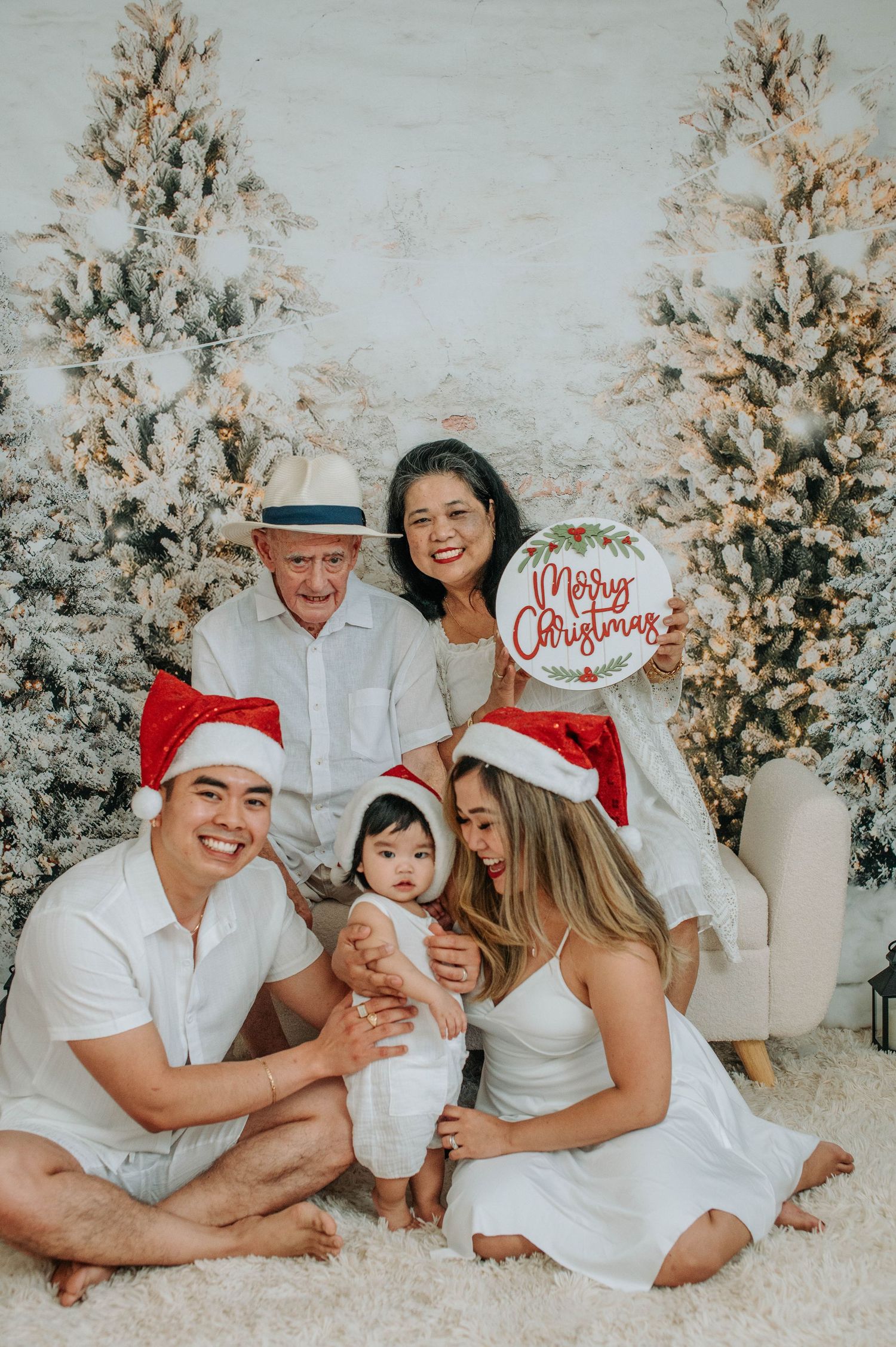 Family wearing white outfits poses together on white carpet with decorated Christmas trees in the background.