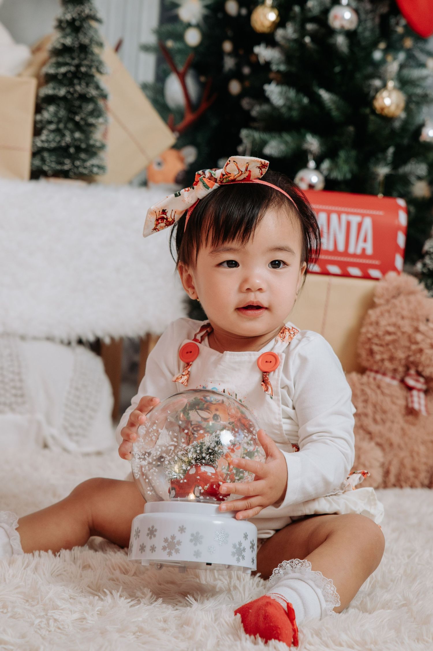 Baby in white dress with floral details sits among Christmas decorations and plush toys on white fur rug.