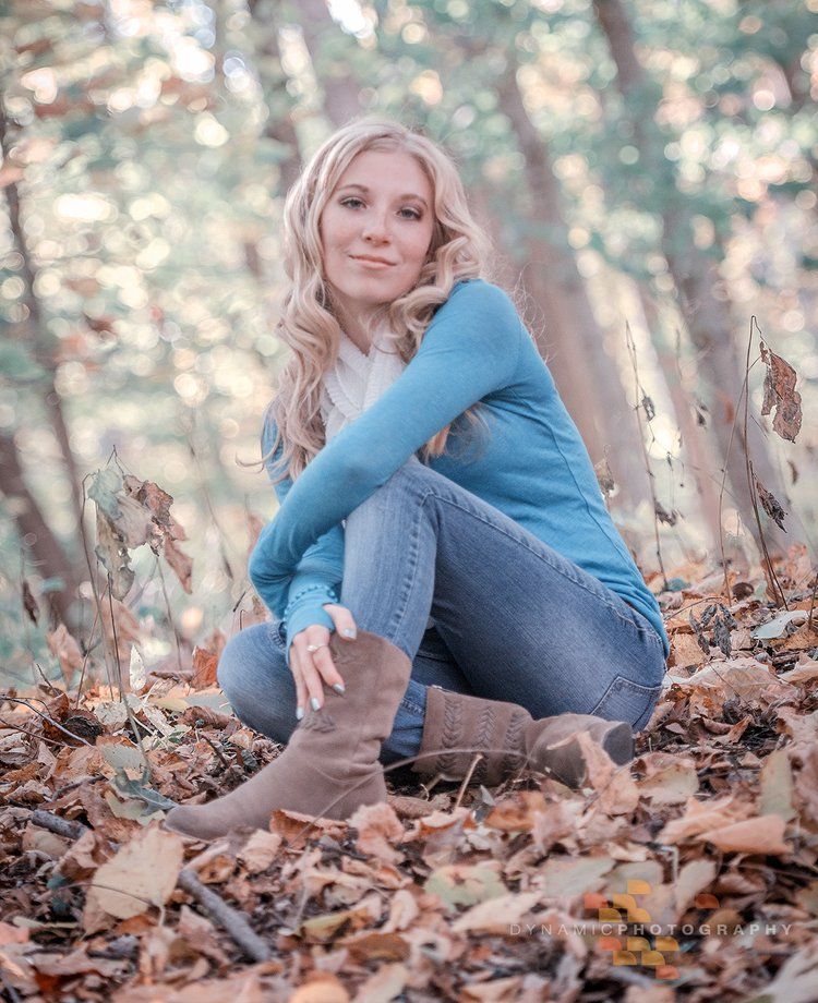 A Thornton Colorado Senior in blue sweater and jeans sits among autumn leaves in a dreamy outdoor portrait setting.