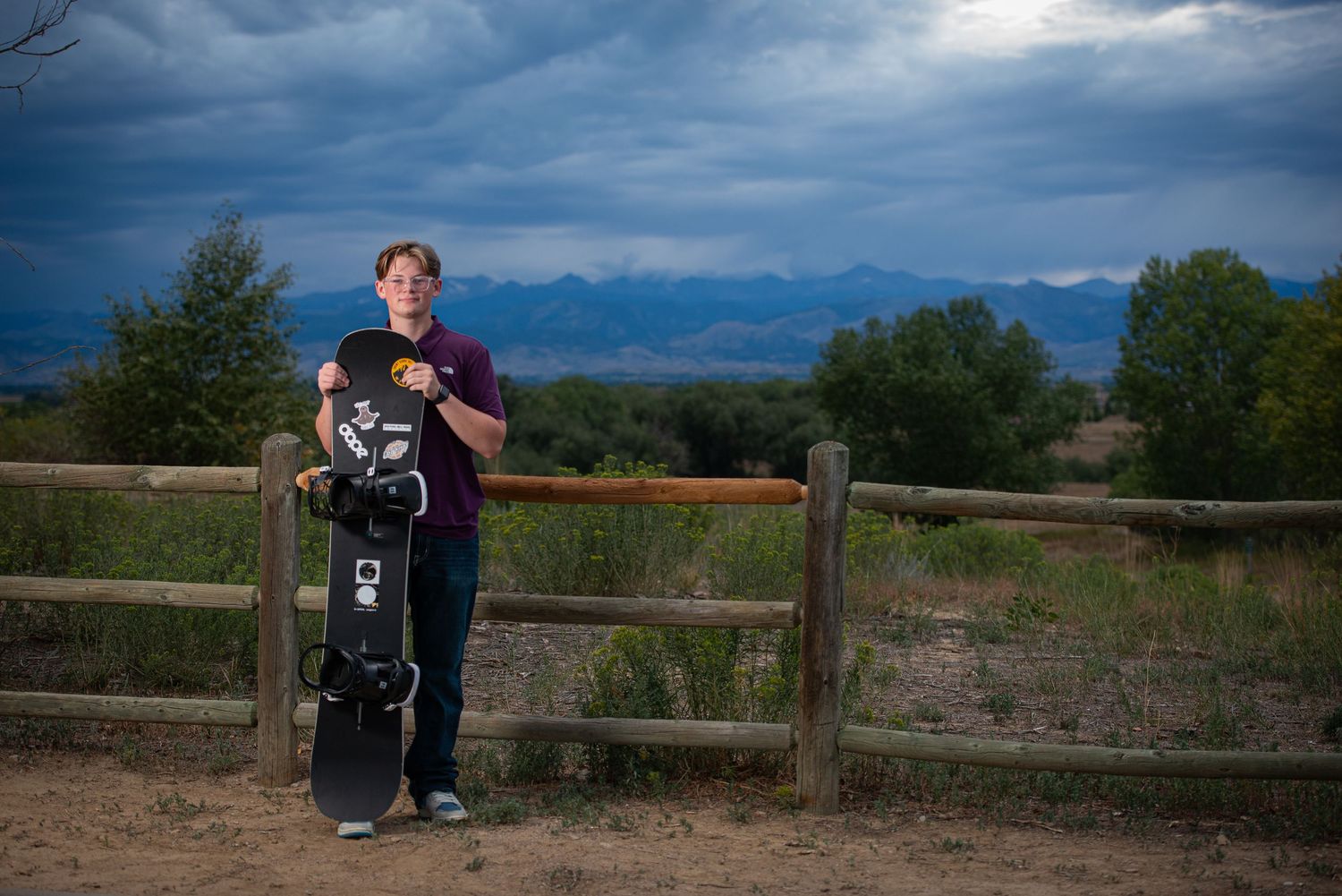 A Broomfield Senior sits with a panoramic view of a wooden fence stretches across rolling hills under dramatic cloudy skies at sunset.