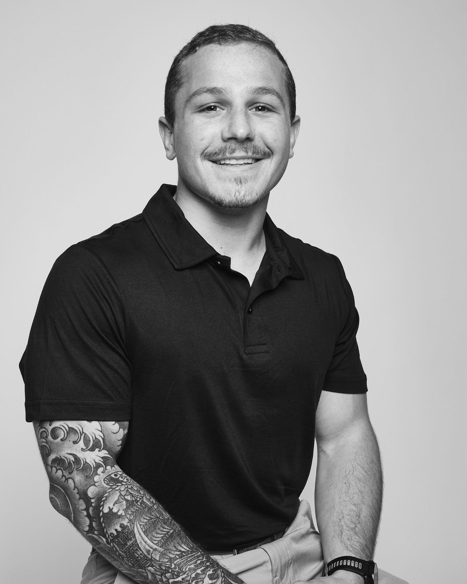 Smiling young man with sleeve tattoo wearing black polo shirt in black and white portrait photo.