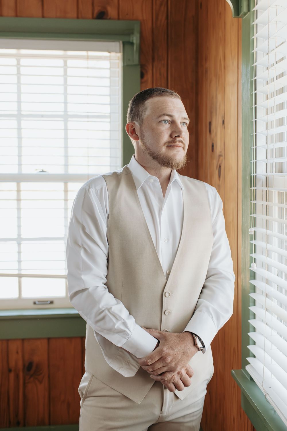 Groom in light beige suit getting ready in wood-paneled room with natural light.