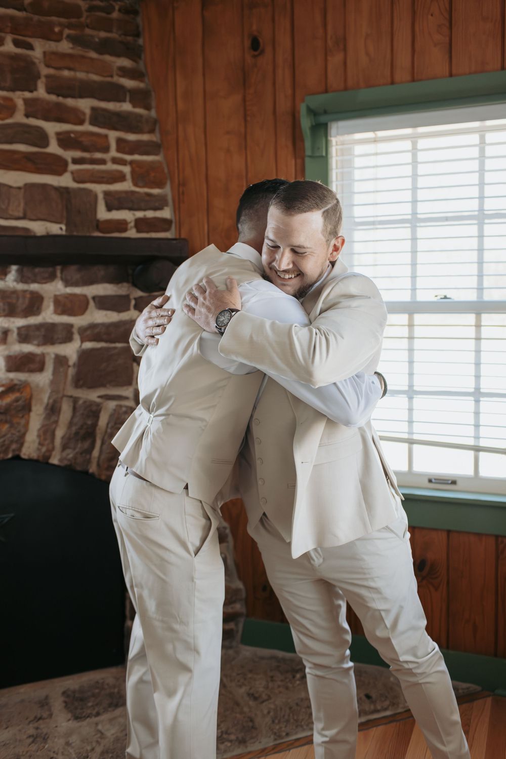 Two groomsmen in cream colored suits playfully dab together in a rustic indoor setting.