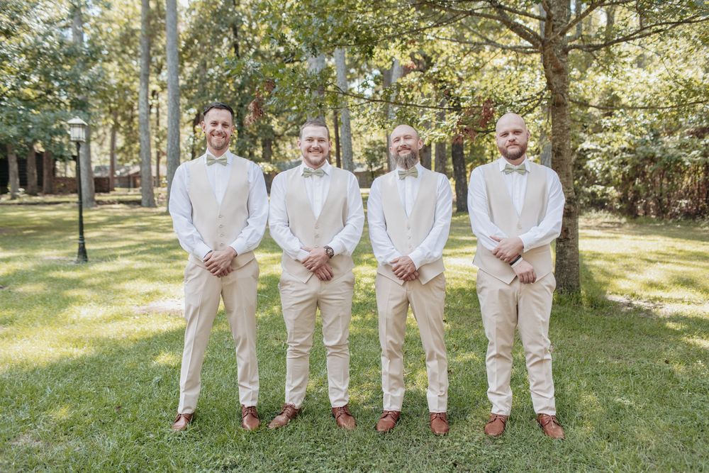 Wedding party members in cream suits pose together on a sunny lawn with trees in background.