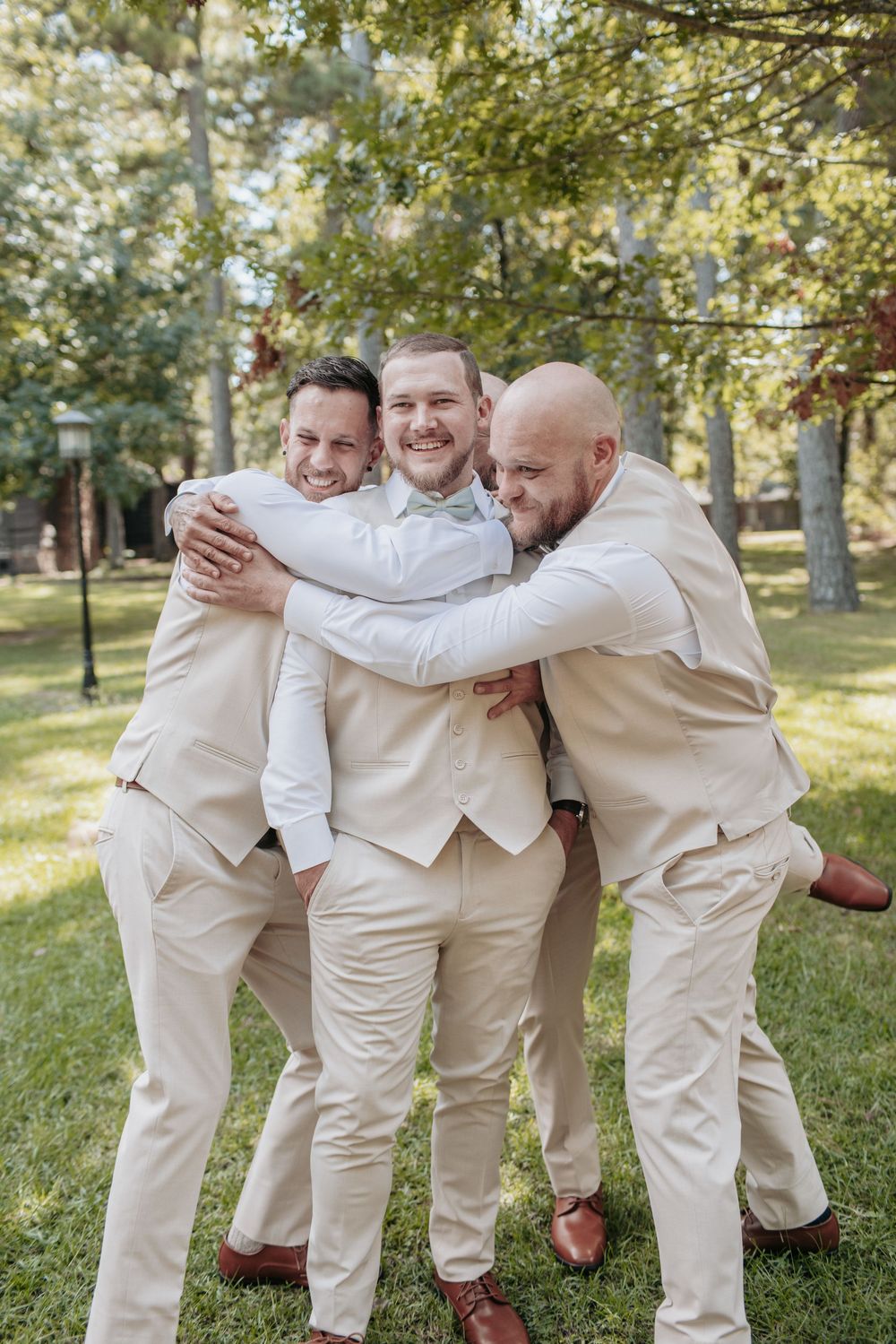 Groomsmen in cream suits playfully pose and hug on a sunny lawn.