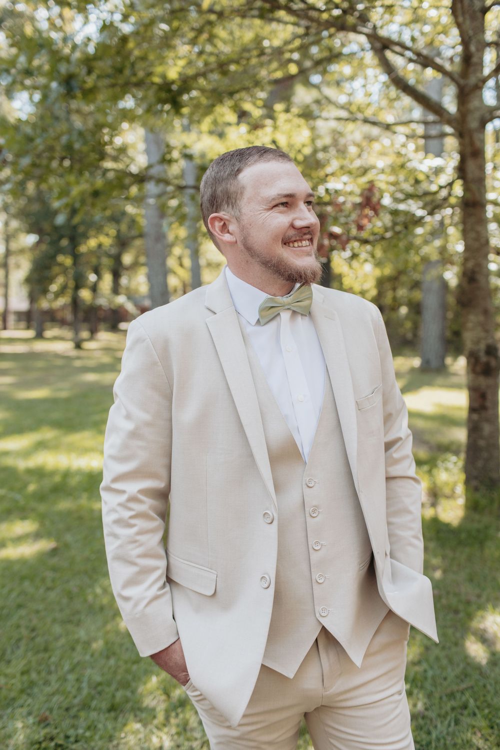Groom smiling and adjusting his cream colored suit jacket outdoors.