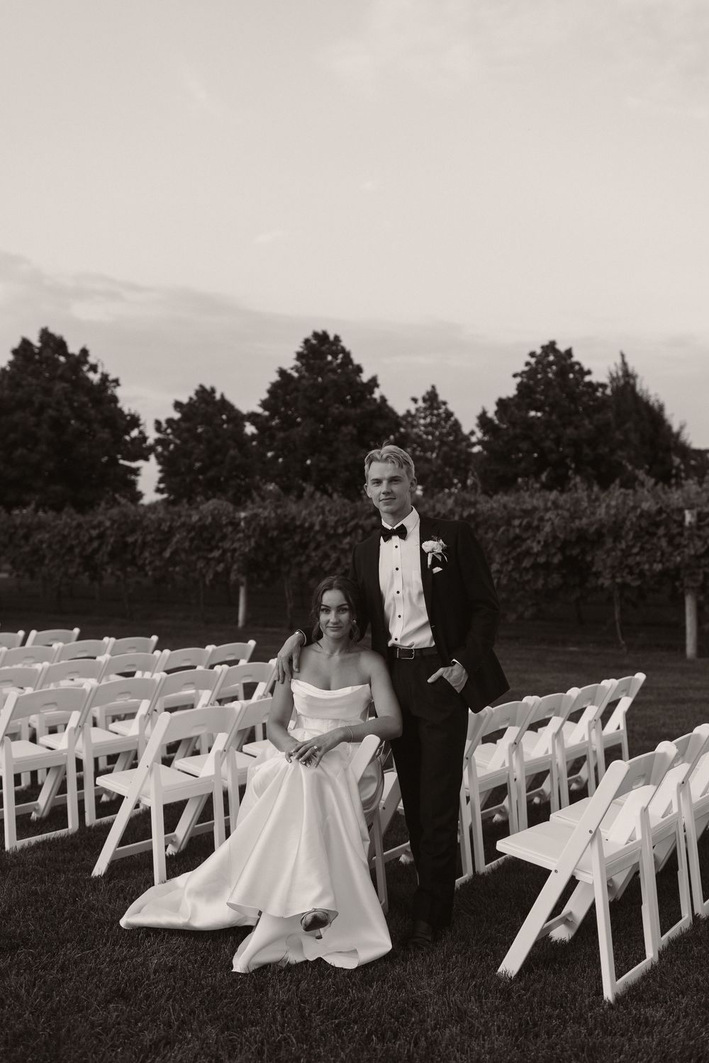 Couple in wedding attire sitting among white ceremony chairs in black and white.