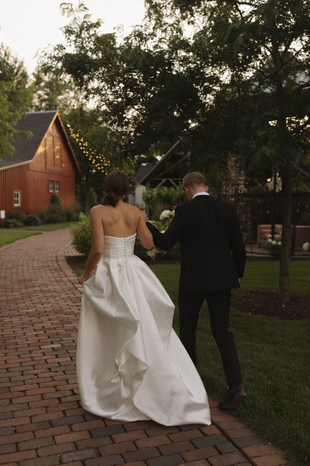 Wedding couple walking along a brick pathway near a rustic red barn at dusk.