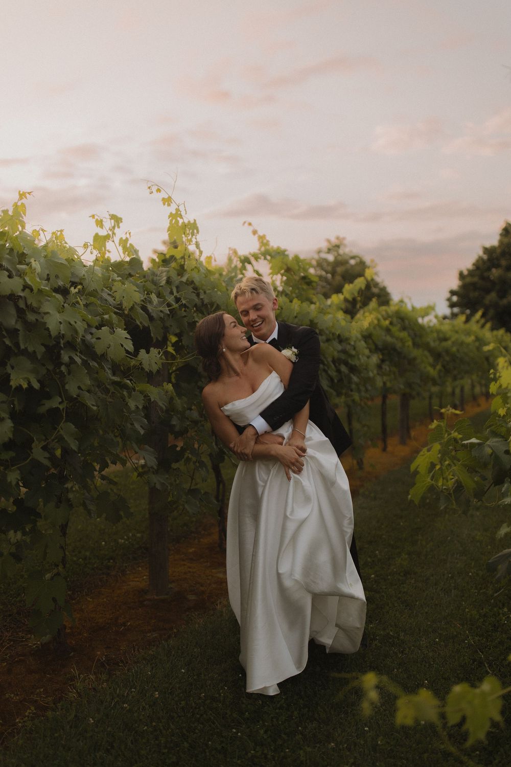 Couple in wedding attire sharing a romantic moment between grapevines.