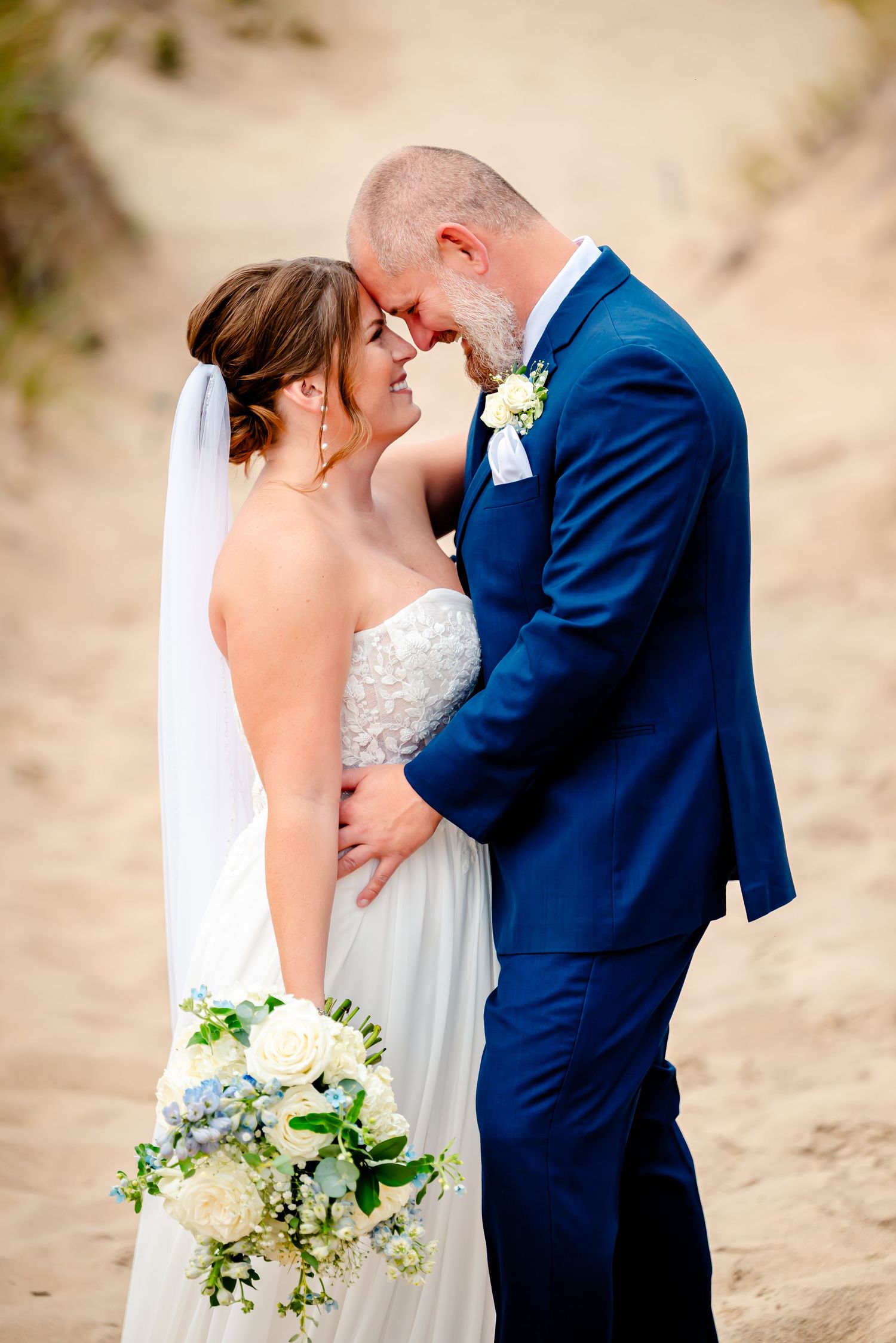 A romantic wedding couple shares an intimate moment on a sandy beach with a white and blue floral bouquet.