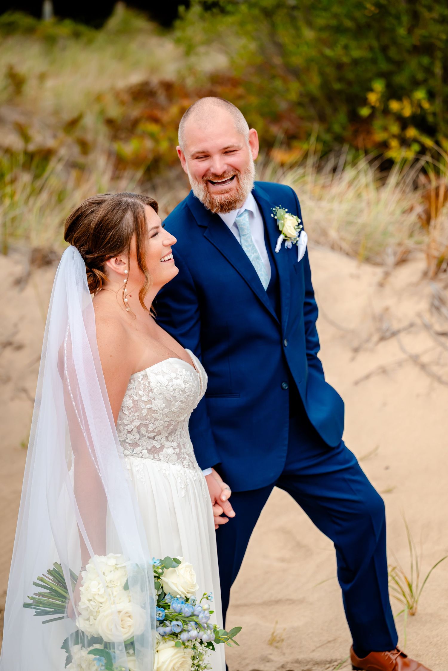 A happy couple shares a joyful moment on a sandy beach during their wedding, with the bride wearing white and groom in navy blue.