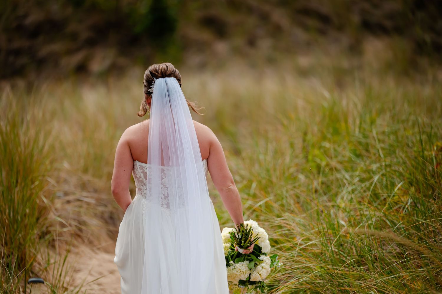 A bride in a white wedding gown and veil stands in a field of tall grass at sunset.