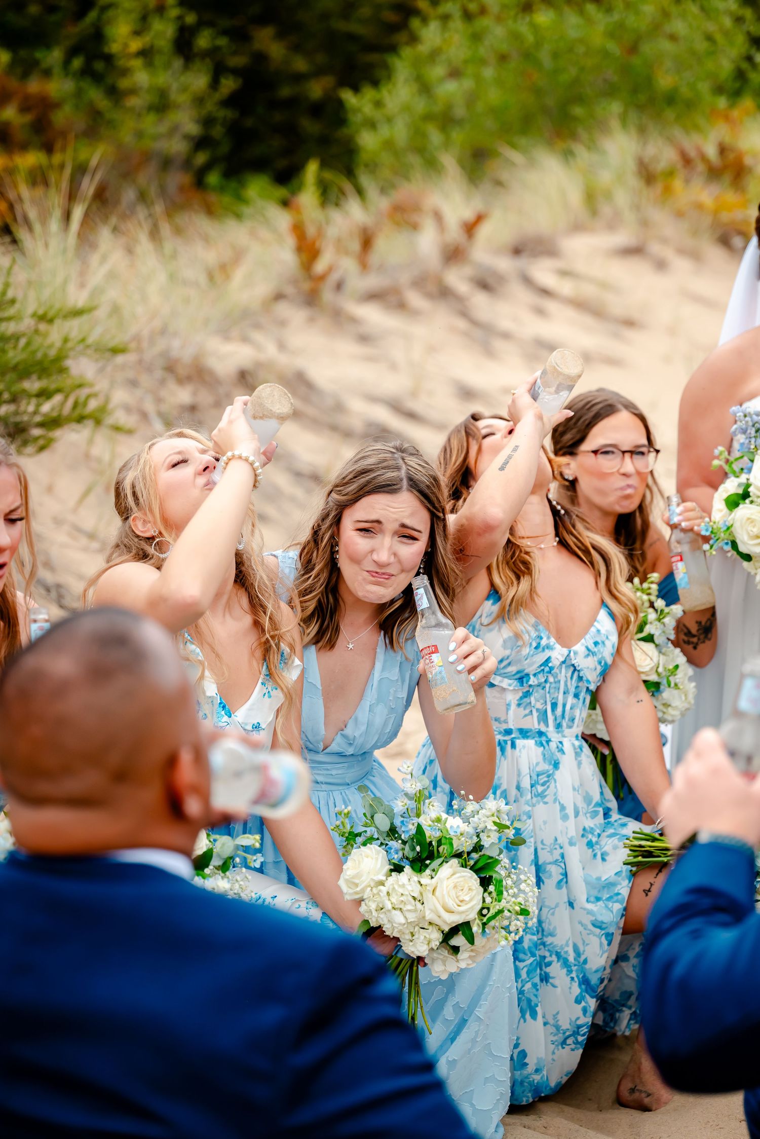 Wedding party in light blue dresses and navy suits laughs together on beach dunes.