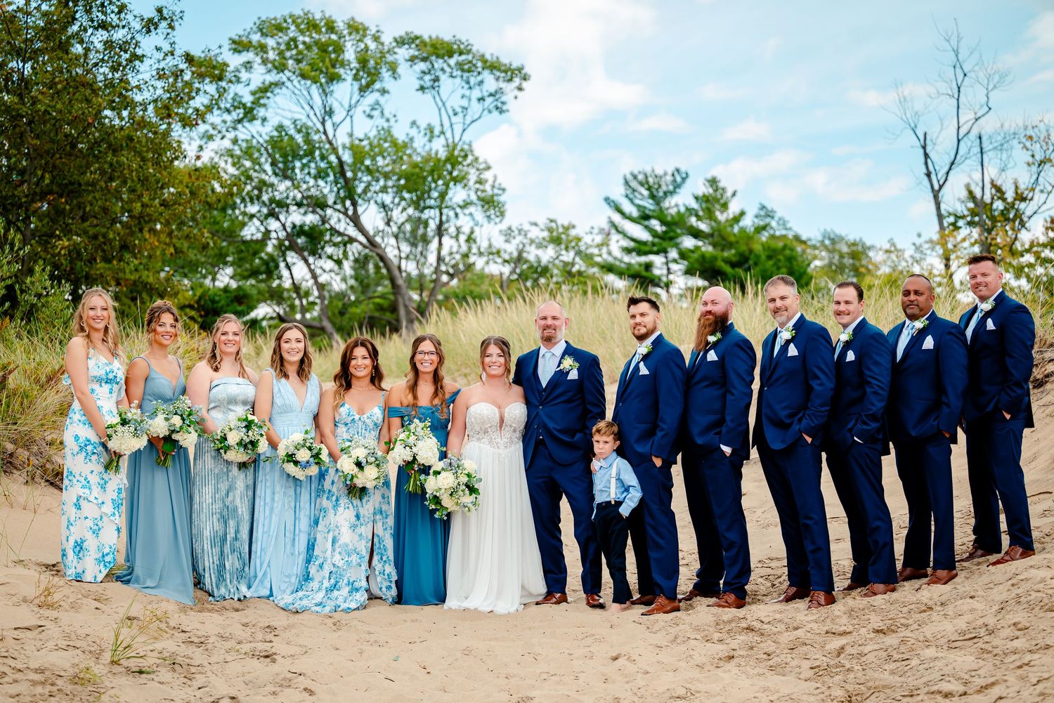 Wedding party in light blue dresses and navy suits pose together on a sandy beach with ocean views.