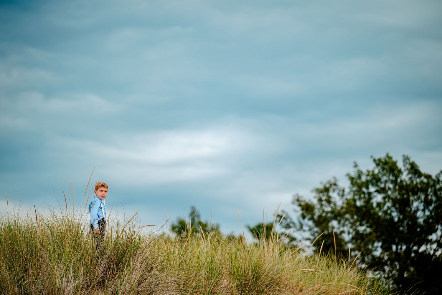 A row of groomsmen in navy blue suits stand against a cloudy sky and grassy landscape.