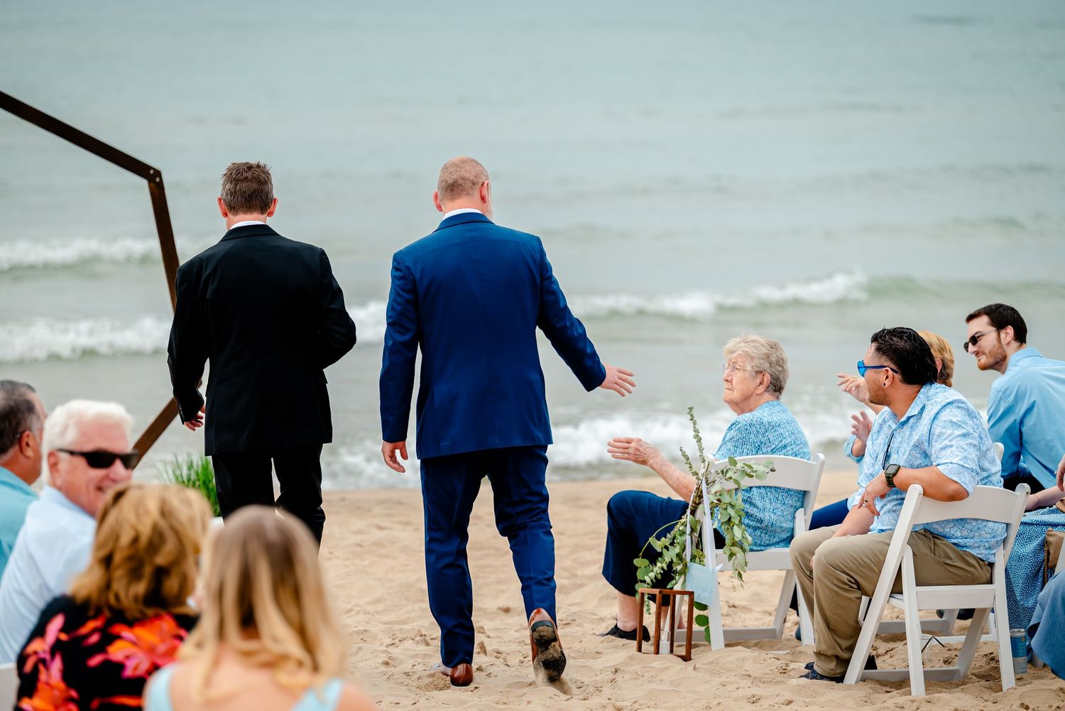 People sitting in white chairs on a beach during a seaside event with waves in the background.