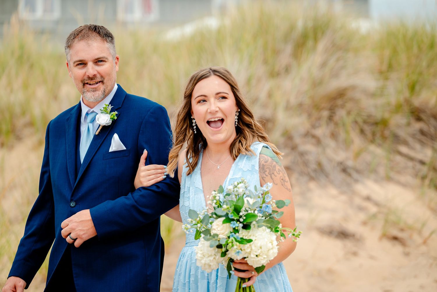 Newlywed couple in formal attire poses for romantic photos on a sandy beach with grass in the background.