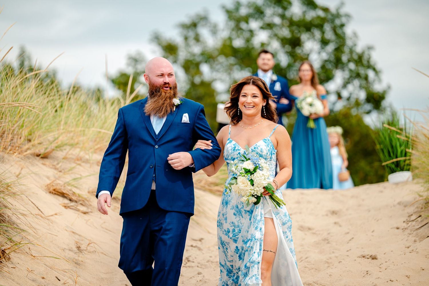 Wedding party in blue and floral dresses walking along coastal sand dunes.