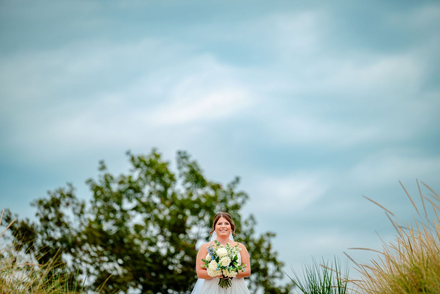 A person in a white dress holds a floral bouquet against a cloudy blue sky with green foliage in the background.
