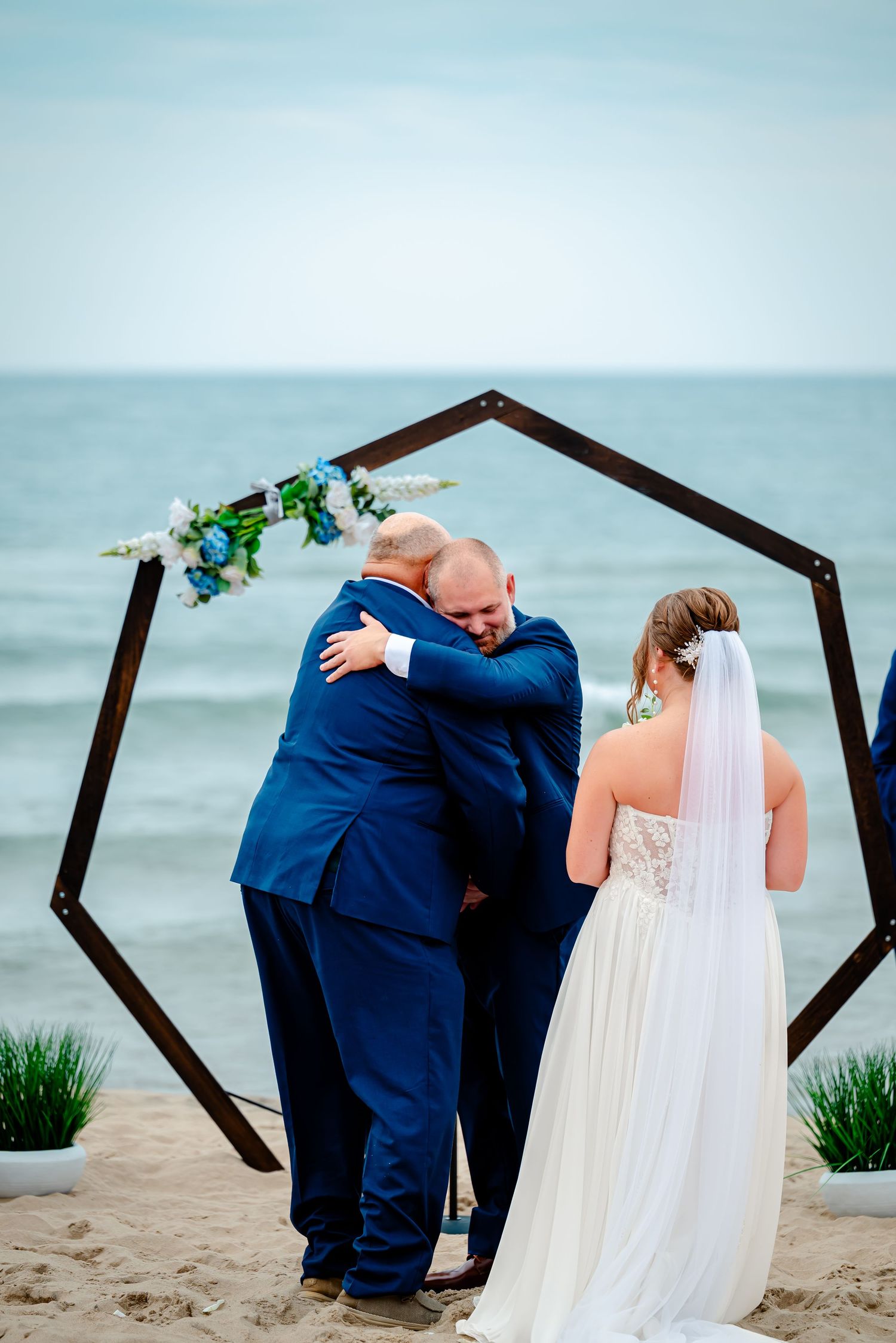 A wedding couple shares an intimate moment under a geometric arch on a sandy beach with ocean views.