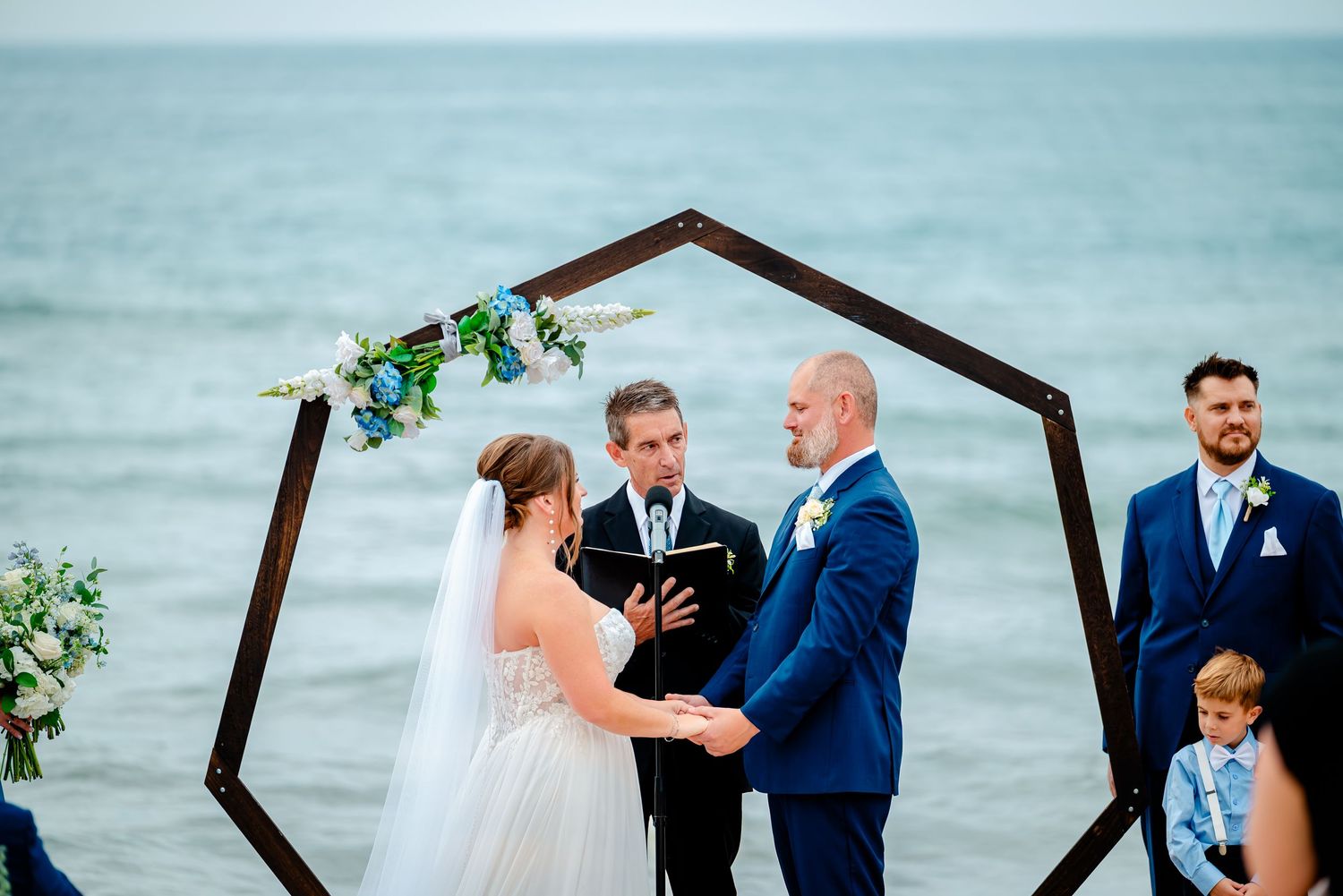 Wedding ceremony setup on beach with wooden hexagonal arch and white chairs overlooking the ocean.