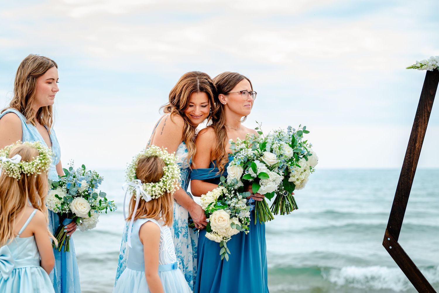 Wedding party members in light blue dresses and navy suits stand together against beach backdrop.