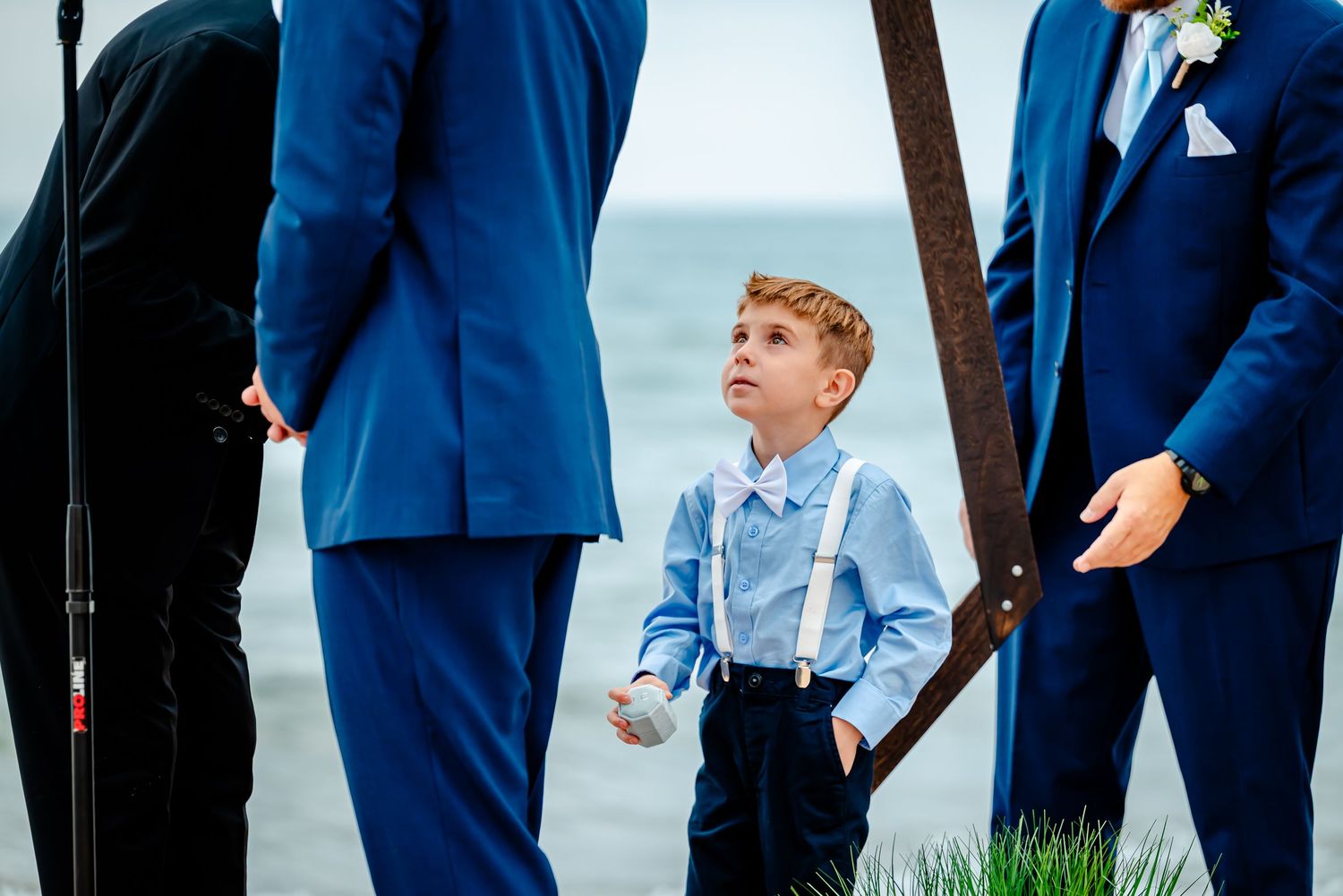 Wedding party members in navy suits stand together on a grassy hill with an overcast sky backdrop.
