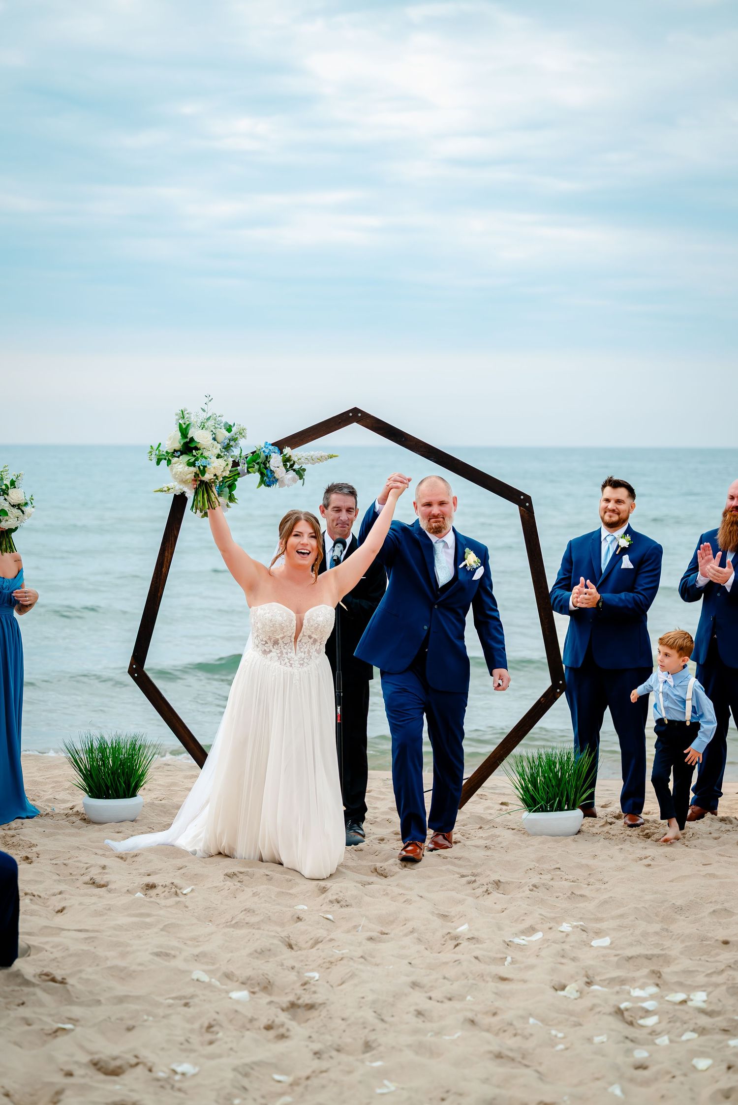 Wedding party poses on beach with geometric arch and ocean backdrop.