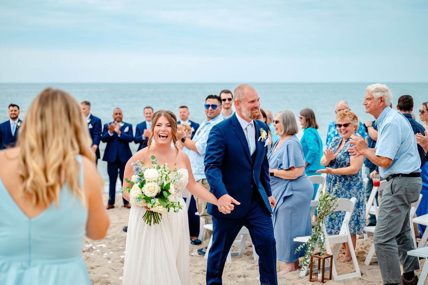 Wedding guests gather on a sunny beach during a seaside ceremony with blue ocean views in the background.